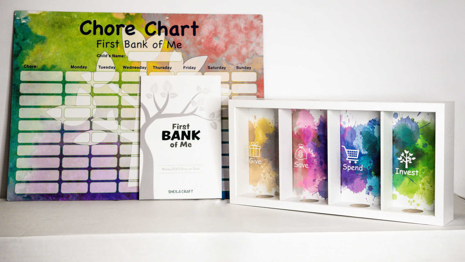 'First Bank of Me' bundle showcasing a watercolor chore chart and a white book cover with a grey tree, along with a piggy bank that has four designated slots for managing money.