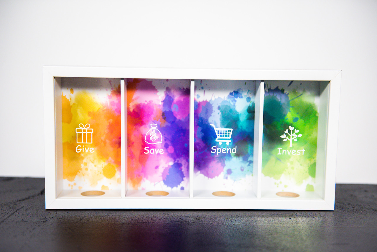 Bank of Me' piggy bank showcasing a clear front window displaying four separate slots for Give, Save, Spend, and Invest, designed to help children learn about saving.