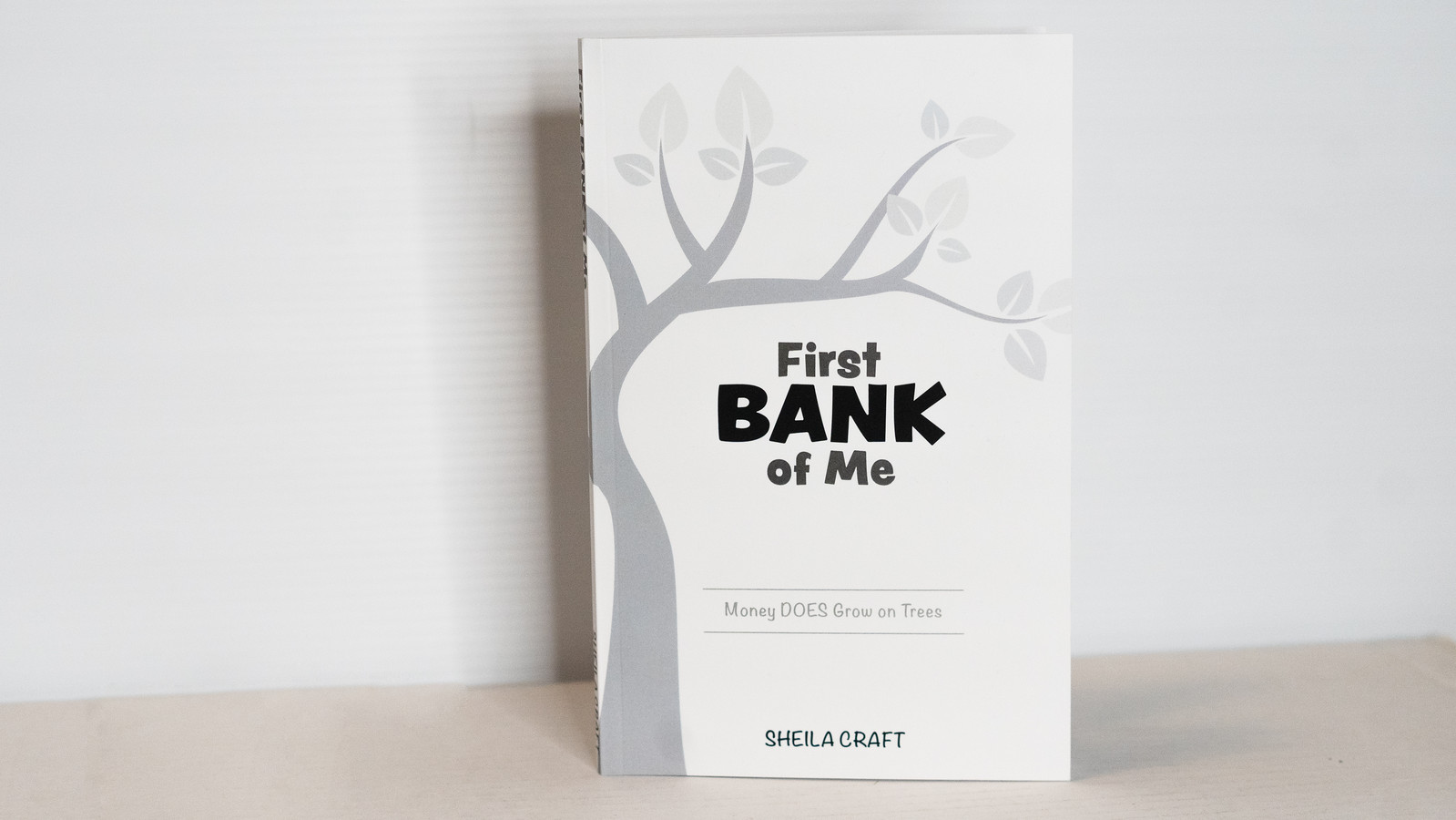'First Bank of Me' book cover, featuring a clean white background with a grey tree and the title, along with the quote 'Money does grow on trees.'